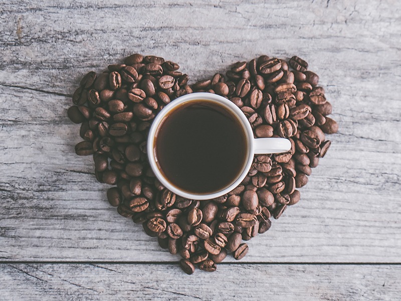 Bird's eye view of coffee beans arranged in the shape of a heart. In the centre of the heart is a mug filled with coffee